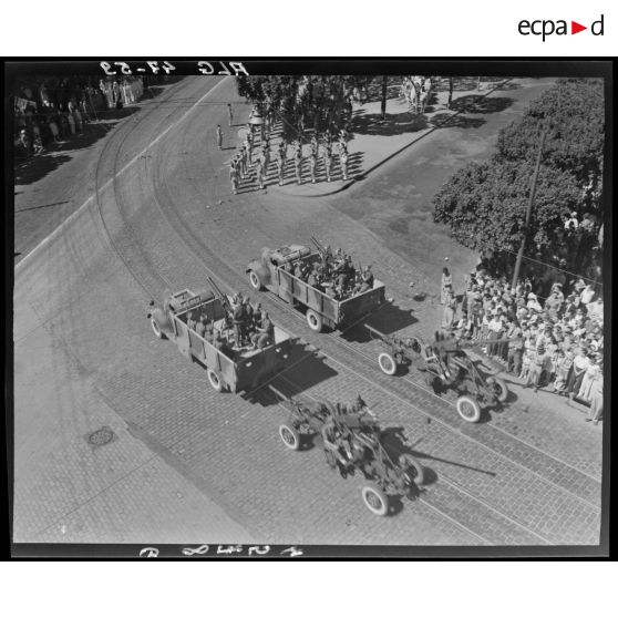 Plan en plongée d'un défilé de canons antiaériens, sous le regard de la population civile, à Alger.