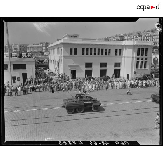 Vue en contre-plongée d'un défilé de véhicules blindés, sous le regard d'autorités civiles et militaires.