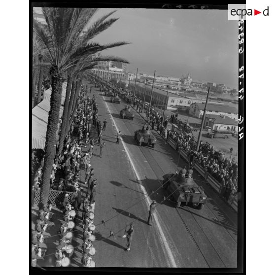 Plan en plongée d'un défilé motorisé, sous le regard d'autorités civiles, militaires et de la population civile.