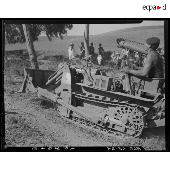 Un militaire déboise un terrain avec un bulldozer équipé d'une tarière, sous le regard de la population civile.