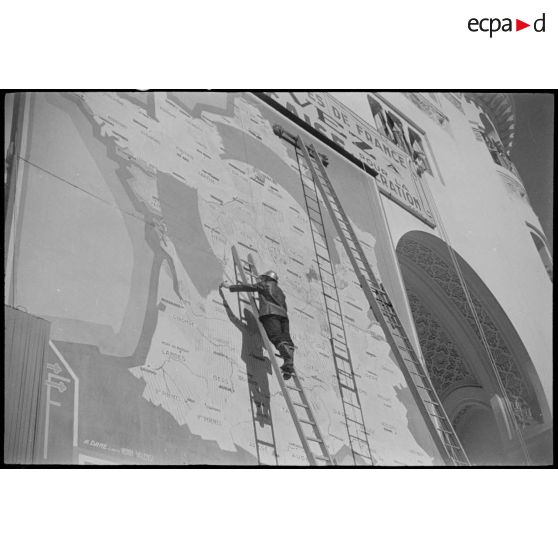 Un sapeur-pompier juché sur une échelle peint sur la carte de France, affichée sur une façade, à la suite de la libération du 8 mai 1945, à Alger.