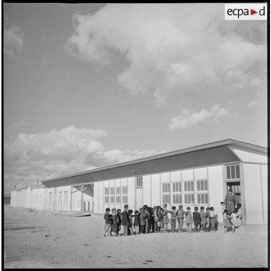 Vue d'une école. Devant l'entrée d'une salle de classe, des élèves patientent en rang avant de rentrer. [légende d'origine]