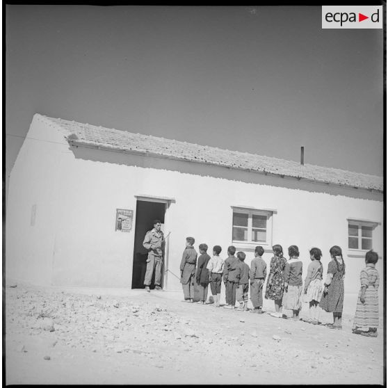 Vue de l'entrée d'une salle de classe. Sur le pas de porte, un instituteur parle à ses élèves qui attendent en rang devant l'entrée de la salle. [légende d'origine]