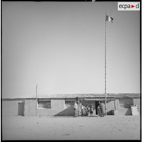 Vue d'ensemble d'une école. Des écoliers attendent en rang devant l'entrée de leur salle de classe. [légende d'origine]