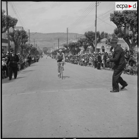 Course cycliste à Saïda. Un coureur cycliste pendant la course. De part et d'autre de la voie, la population venue en nombre observe la course. [légende d'origine]