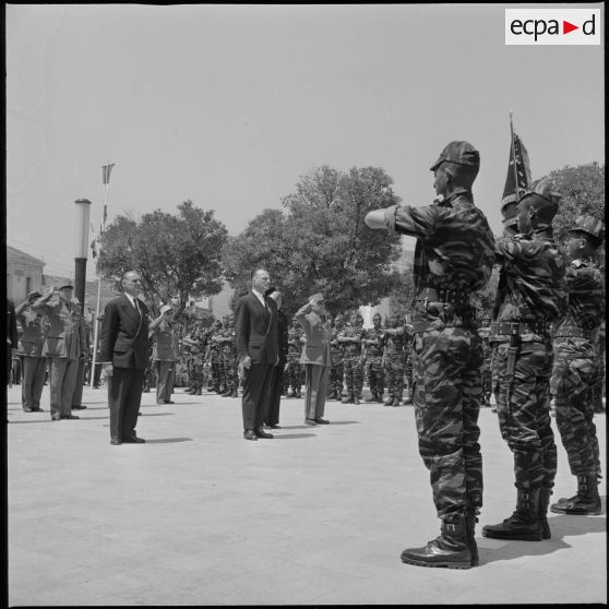 Des membres du commando "George " présentent les armes face aux autorités civiles et militaires rassemblées devant le monument aux morts de Saïda. Au centre, on reconnaît le Délégué général du gouvernement en Algérie Paul Delouvrier. [légende d'origine]