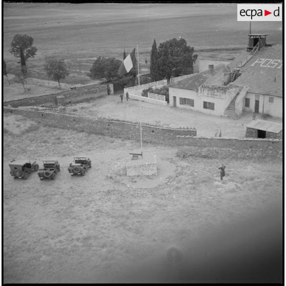 Vue aérienne du poste de Tircine. Vue du mat, du drapeau français et de trois jeeps en stationnement devant le poste. [légende d'origine]