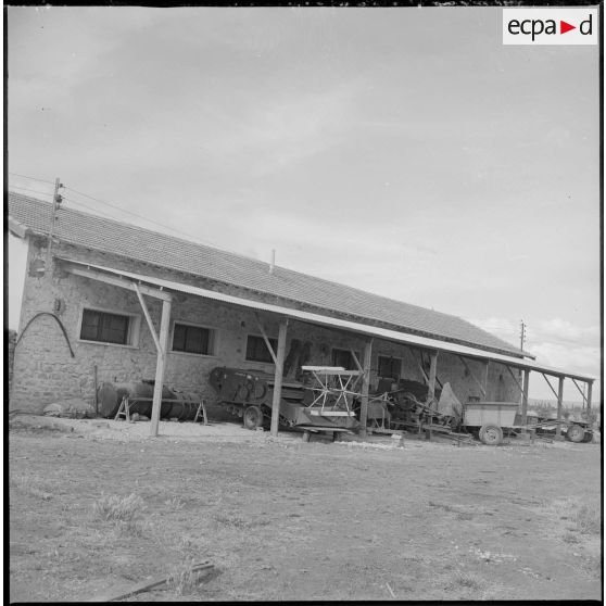 Vue d'un corps de ferme et de machines agricoles stationnées sous un auvent. [légende d'origine]