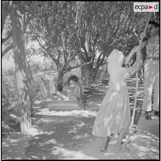 Au Camp des chasseurs du 8e RIM, une jeune fille fait de la balançoire tandis qu'une autre s'apprête à monter à la corde sous la surveillance d'un soldat. [légende d'origine]