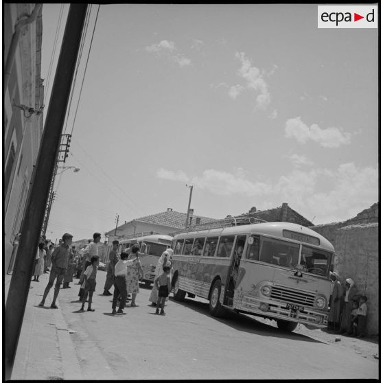 En ville, deux bus venus récupérer des enfants stationnent en attendant le départ pour le Camp des chasseurs du 8e RIM. Plusieurs enfants accompagnés de leurs parents sont visibles devant les bus. [légende d'origine]