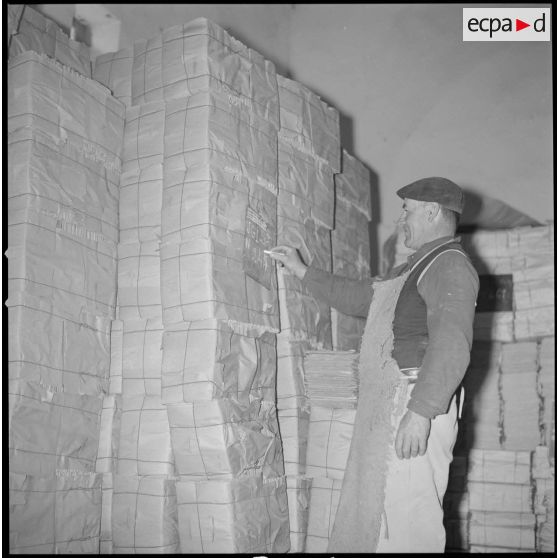 [Papeterie d'Aïn-el-Hadjar. Un ouvrier écrit à l'aide d'une craie sur une ardoise accrochée à un stock de papier.]