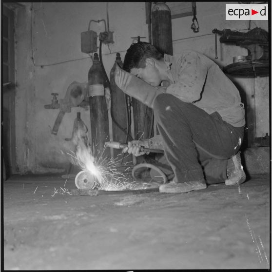 [Papeterie d'Aïn-el-Hadjar. Un ouvrier, accroupi au sol, effectue un travail de soudure.]