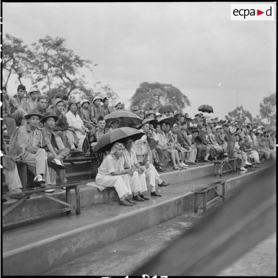 Dans les gradins, sous la pluie, les spectateurs regardent un match opposant l'équipe militaire de France à l'une des équipes vietnamiennes de la sélection du Nord-Vietnam.