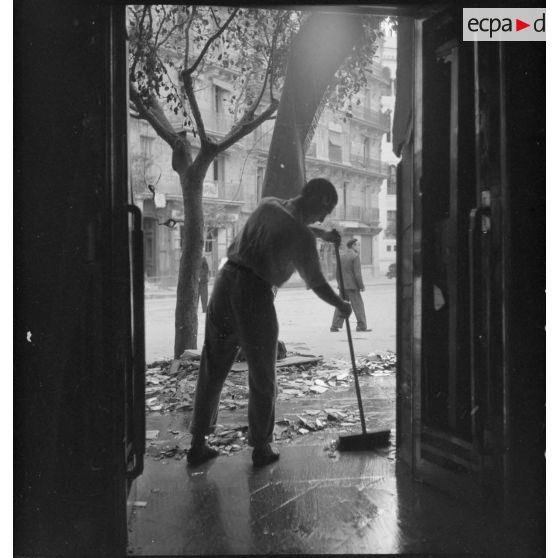 Un habitant balaie le trottoir devant l'entrée d'un immeuble endommagé de la rue d'Isly après le bombardement allemand visant l'aérodrome Maison-Blanche à Alger, dans la nuit du 20 au 21 novembre 1942.