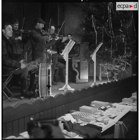 Plan moyen d'un orchestre qui joue sur une scène de théâtre, au premier plan divers cadeaux sont exposés sur des tables.