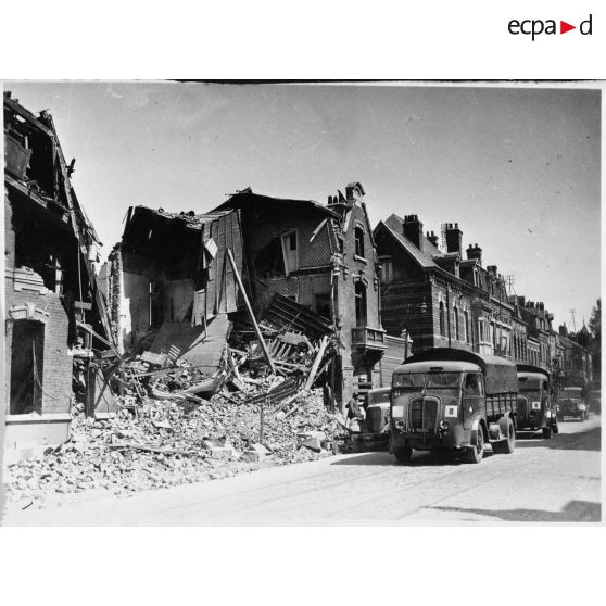 Des camions Renault AGR de la 1re DIM roule de trois quarts face en plan général dans une rue détruite de Valenciennes.