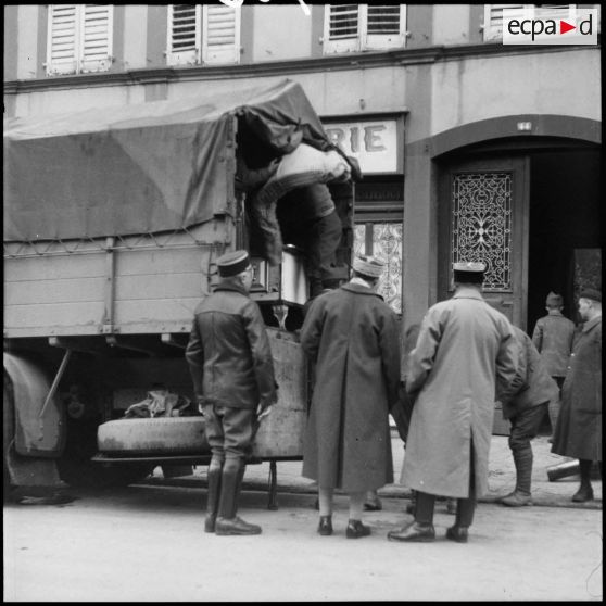 Des soldats de la 3e armée chargent un camion dans une rue de village de la Moselle. Un général les regarde.