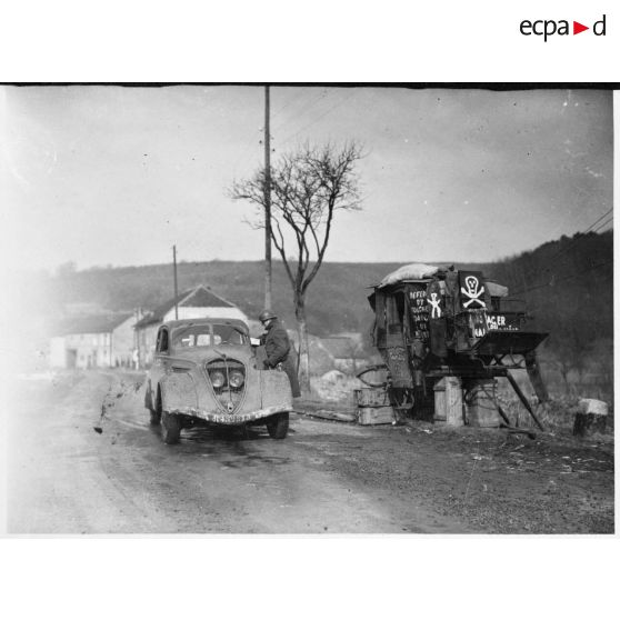 Une Peugeot 202 de la 4e armée est garée devant un vieux fiacre posé sur des caisses de munitions.