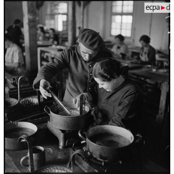 Dans un atelier une ouvrière travaille à la fabrication de piles électriques sous le contrôle d'un militaire.