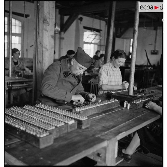 Dans un atelier une ouvrière travaille à la fabrication de piles électriques sous le contrôle d'un militaire.
