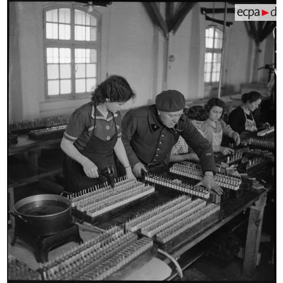 Dans un atelier une ouvrière travaille à la fabrication de piles électriques sous le contrôle d'un militaire.