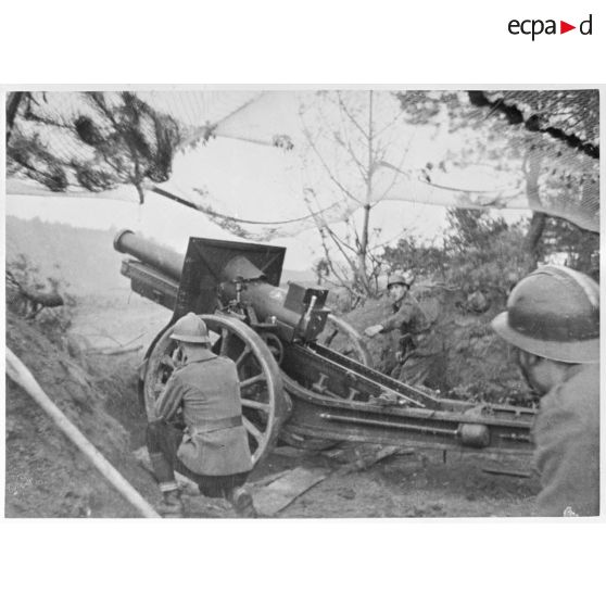 Tir d'une pièce de 155 mm court M1915/17. Cette dernière est photographiée en plan général de trois quarts dos.