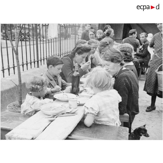 A l'extérieur dans une cour d'école, des femmes et des enfants mangent à table.