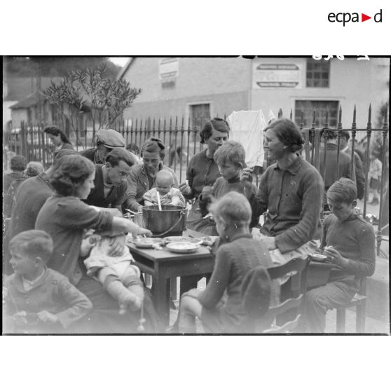 A l'extérieur dans une cour d'école, des femmes et des enfants mangent à table.