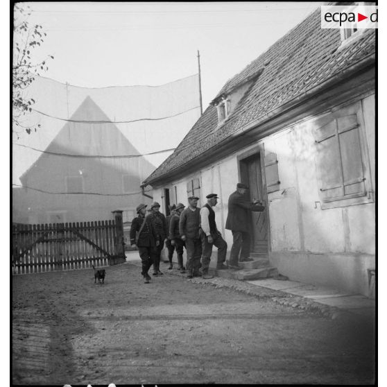 Des soldats et un douanier invités à entrer dans une maison de village par un habitant.