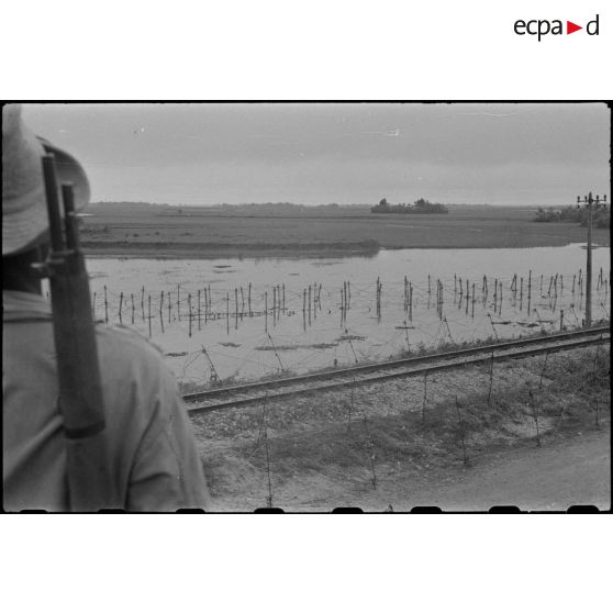Le paysage vu du blockhaus. Au premier plan, une voie ferrée et de dos, un soldat armé.