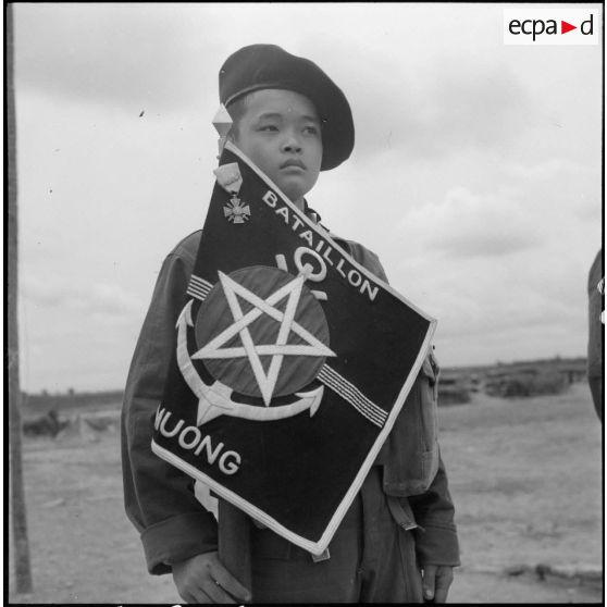 Un tirailleur de 2e classe présente le fanion du 1er bataillon muong, décoré de la croix de guerre des théâtres d'opérations extérieures (TOE), reçue après les combats de Vinh Yen en janvier 1951.