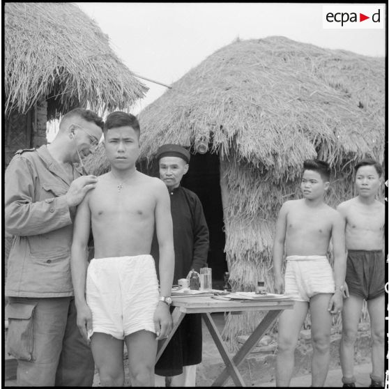Visite médicale lors de l'incorporation des jeunes recrues muong par le médecin-lieutenant, sous le regard du conseiller militaire muong.
