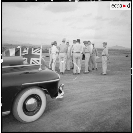 Discussion en groupe des officiels devant la voiture officielle arborant le drapeau français et le drapeau britannique.