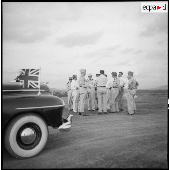 Discussion en groupe des officiels devant la voiture officielle arborant le drapeau français et le drapeau britannique.