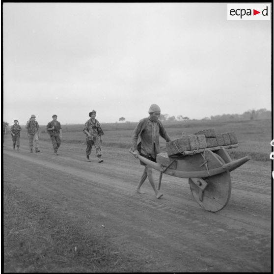 Un villageois transporte des paquets sur une brouette en bois. Des éléments du 8e BCP le suivent.