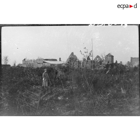 [Soldat posant dans un champ. En fond, village en ruines].<br>