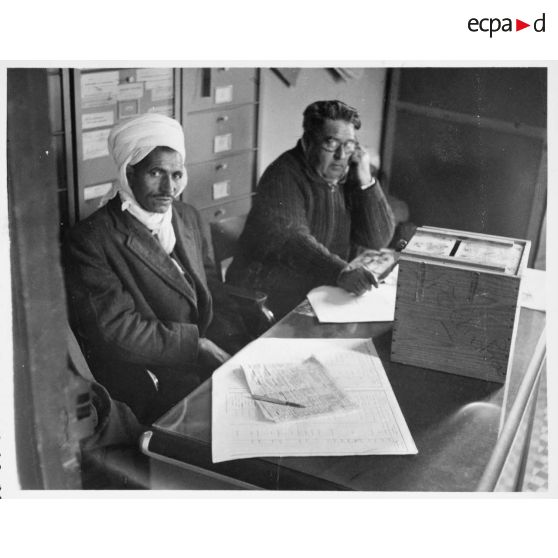 Personnel administratif assis devant une urne servant au vote. [légende d'origine]<br>