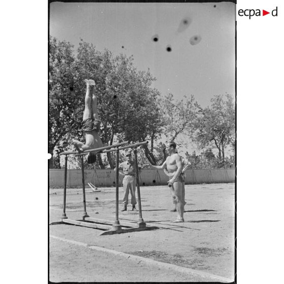 Un jeune engagé militaire s'entraîne aux barres parallèles, en plein air, sous le regard d'autorités militaires.