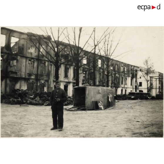 [Un mécanicien allemand pose devant dépôt de réparation. En arrière-plan, des destructions causées par des bombardements. Renversée, une ambulance du service de santé de l'armée de Terre allemande].
