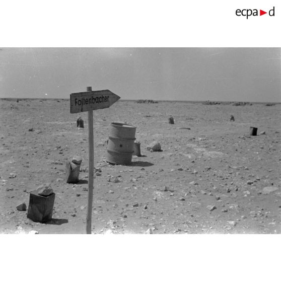 Panneau (flèche) vers la base "Faltenbach", route du désert balisée par de vieux bidons d'essence, de vieux tonneaux au carrefour, à droite, des champs de mines à gauche.