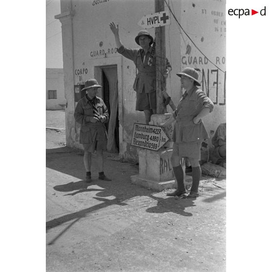 Trois artistes posent devant un ancien poste de garde et un panneau indicateur donnant des indications de distances.