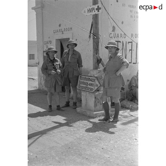 Trois artistes posent devant un ancien poste de garde et un panneau indicateur donnant des indications de distances.