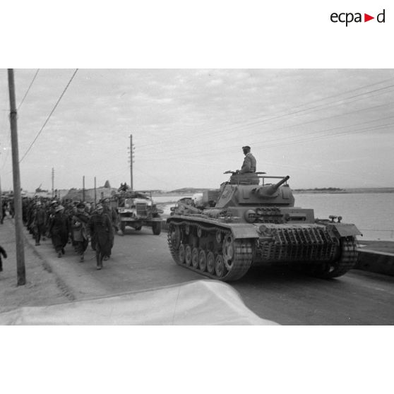 La colonne de prisonniers britanniques qui marche le long de la côte est dépassée par une colonne de véhicules.