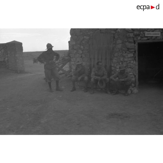 Au sein du fort de Mechili, des soldats italiens, dont trois sont assis et l'un fume, discutent près d'un baraquement.