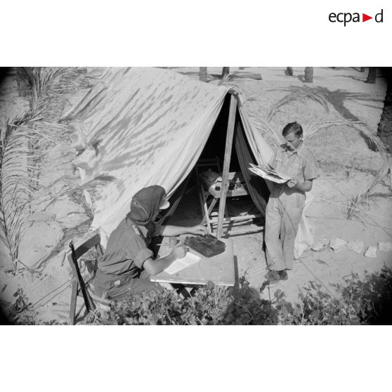 Un Leutnant écrit sur une table, il a recouvert sa casquette d'une moustiquaire, un autre lit près d'une tente.