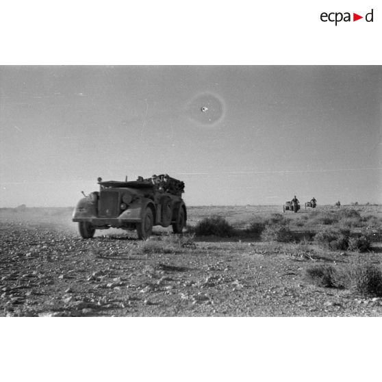 Des voitures Kfz-15 suivies de side-cars roulent dans le désert, l'une des voitures porte un fanion.