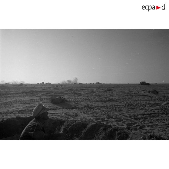 Un soldat abrité dans un trou observe des impacts, au second plan, des véhicules allemands disséminés dans le désert.