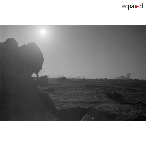 Un soldat abrité dans un trou observe des impacts, au second plan, des véhicules allemands disséminés dans le désert.