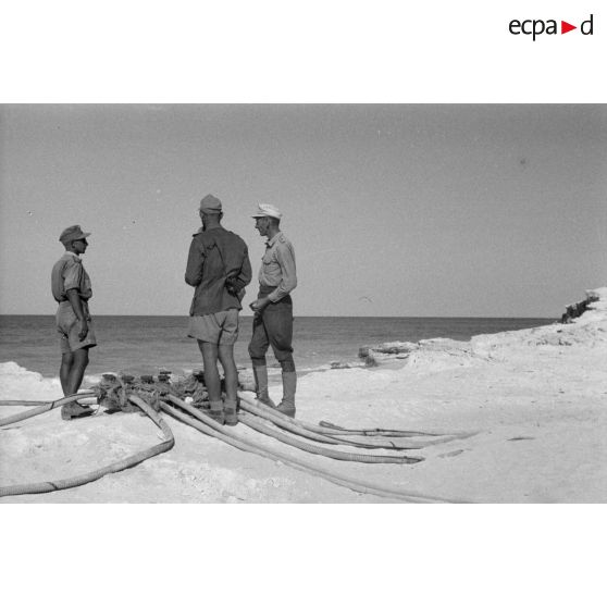 Un groupe d'officiers et de sous-officiers près des tuyaux sur la plage.