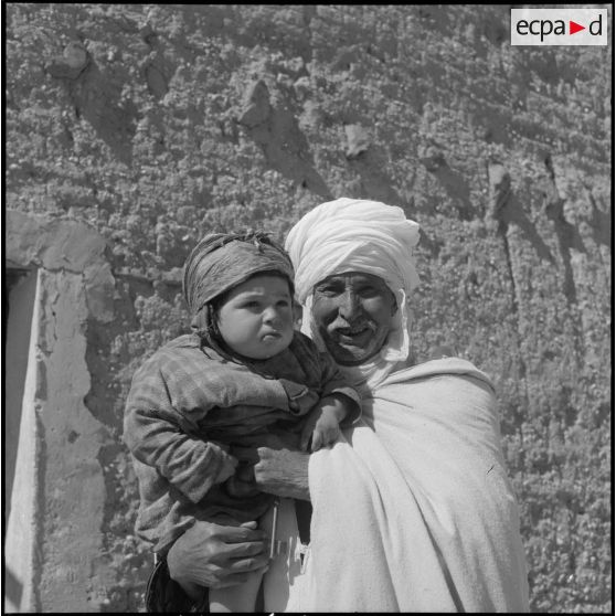 Portrait d'un homme kabyle âgé portant un enfant devant une mechta à M'Chounene dans les Aurès-Nementcha.